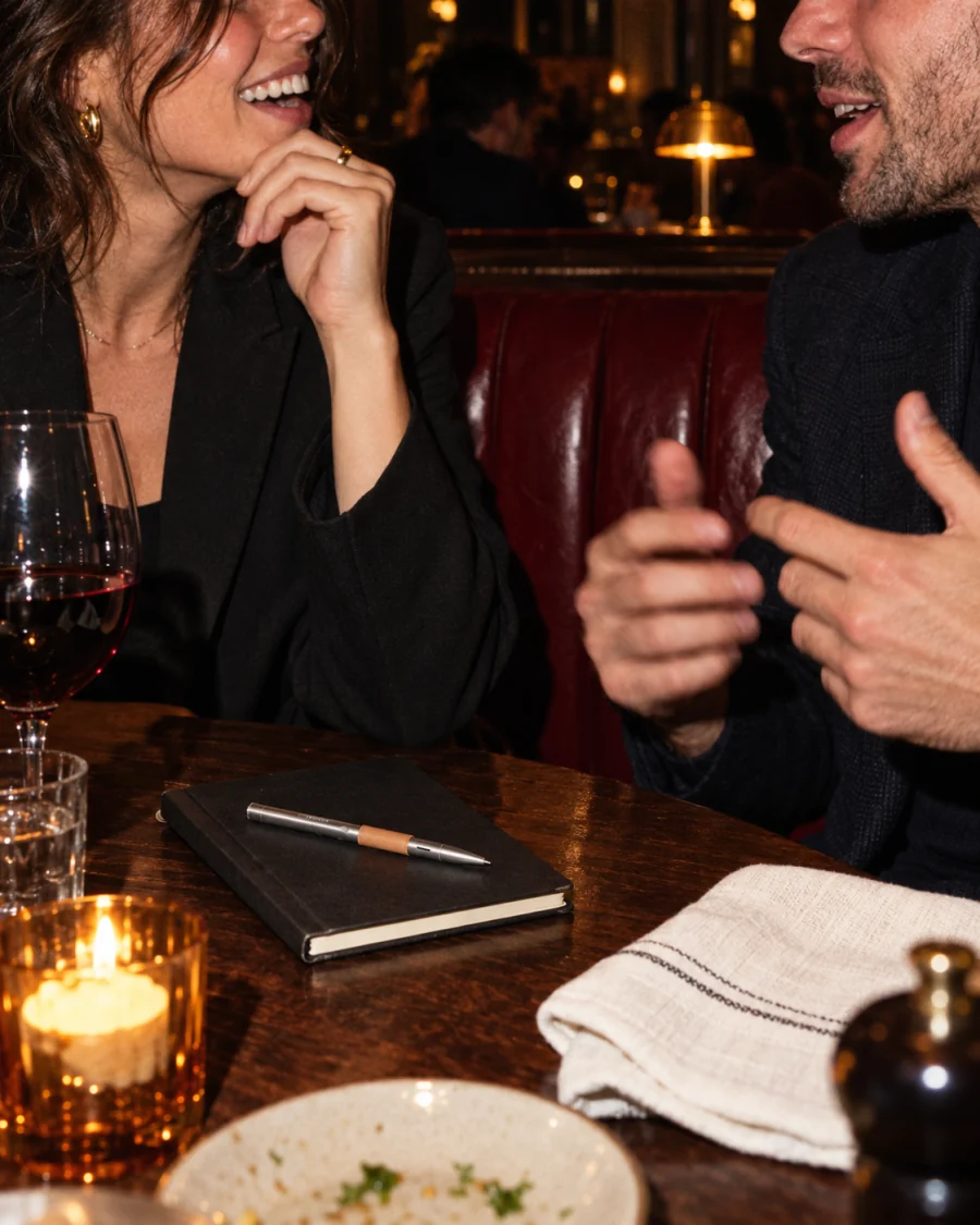 Two people laughing over dinner, the pen resting on a closed notebook.